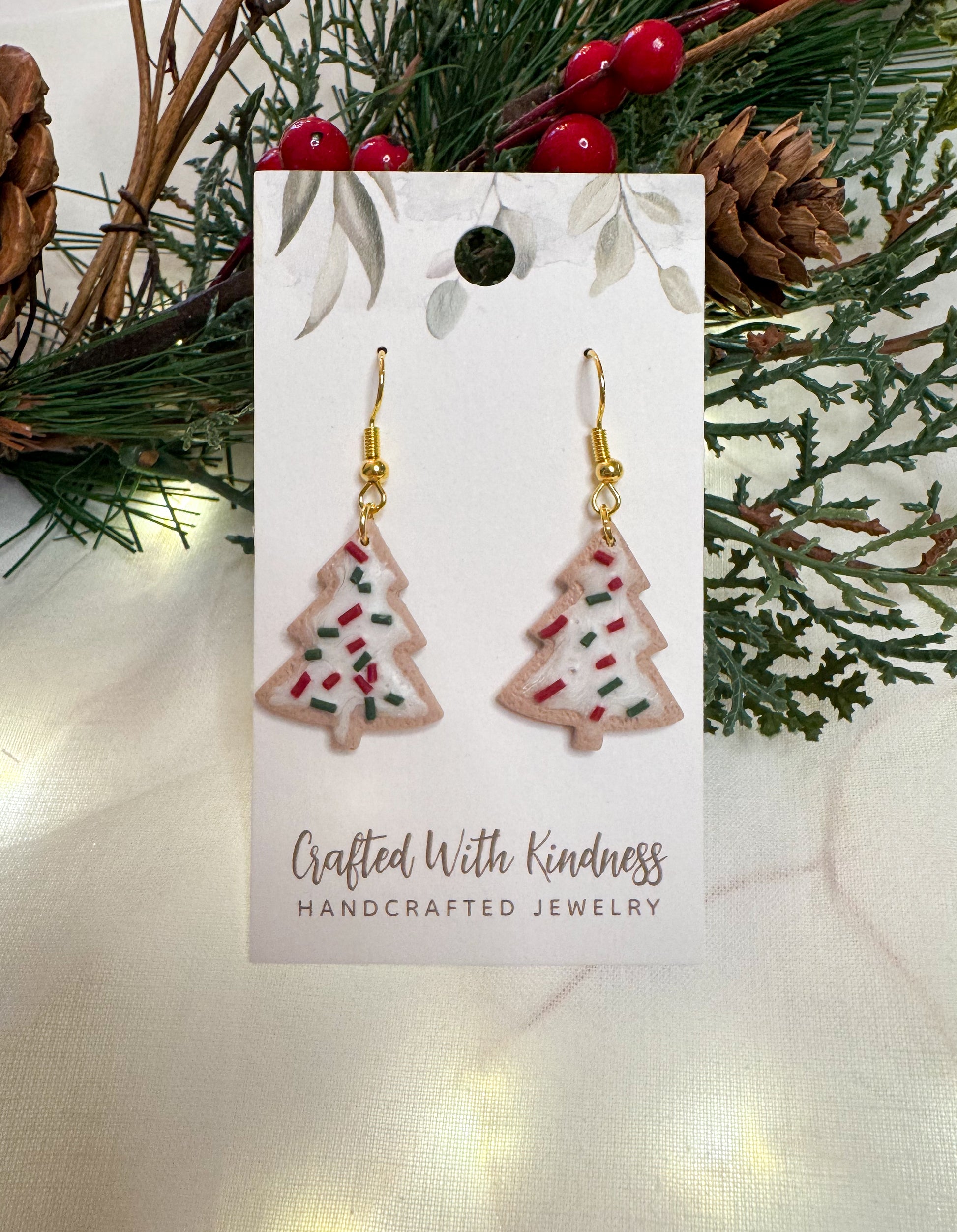 Christmas cookie earrings in the shape of Christmas trees with white frosting and green and red sprinkles.