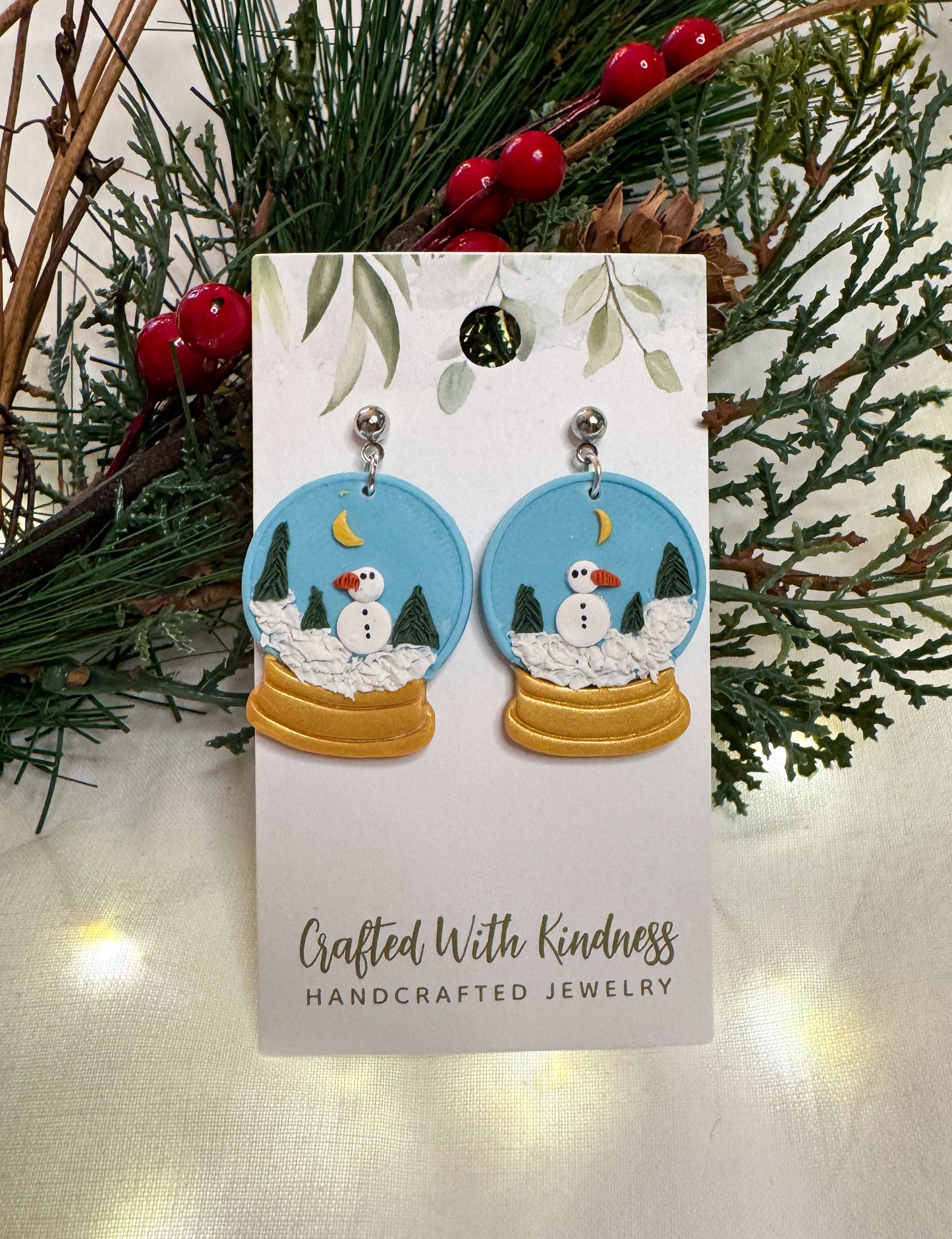 Snow globe inspired earrings made out of polymer clay with a blue sky background, snowy terrain, and a snowman. 